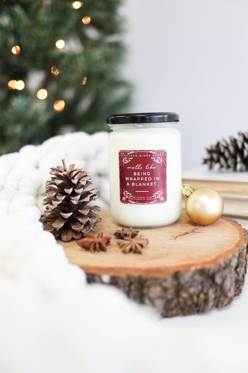 Being Wrapped In A Blanket Candle displayed on a wooden slice with pinecones, cozy ambiance for the holidays.
