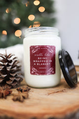 Being Wrapped In A Blanket Candle in a glass jar with holiday decor and pinecones.