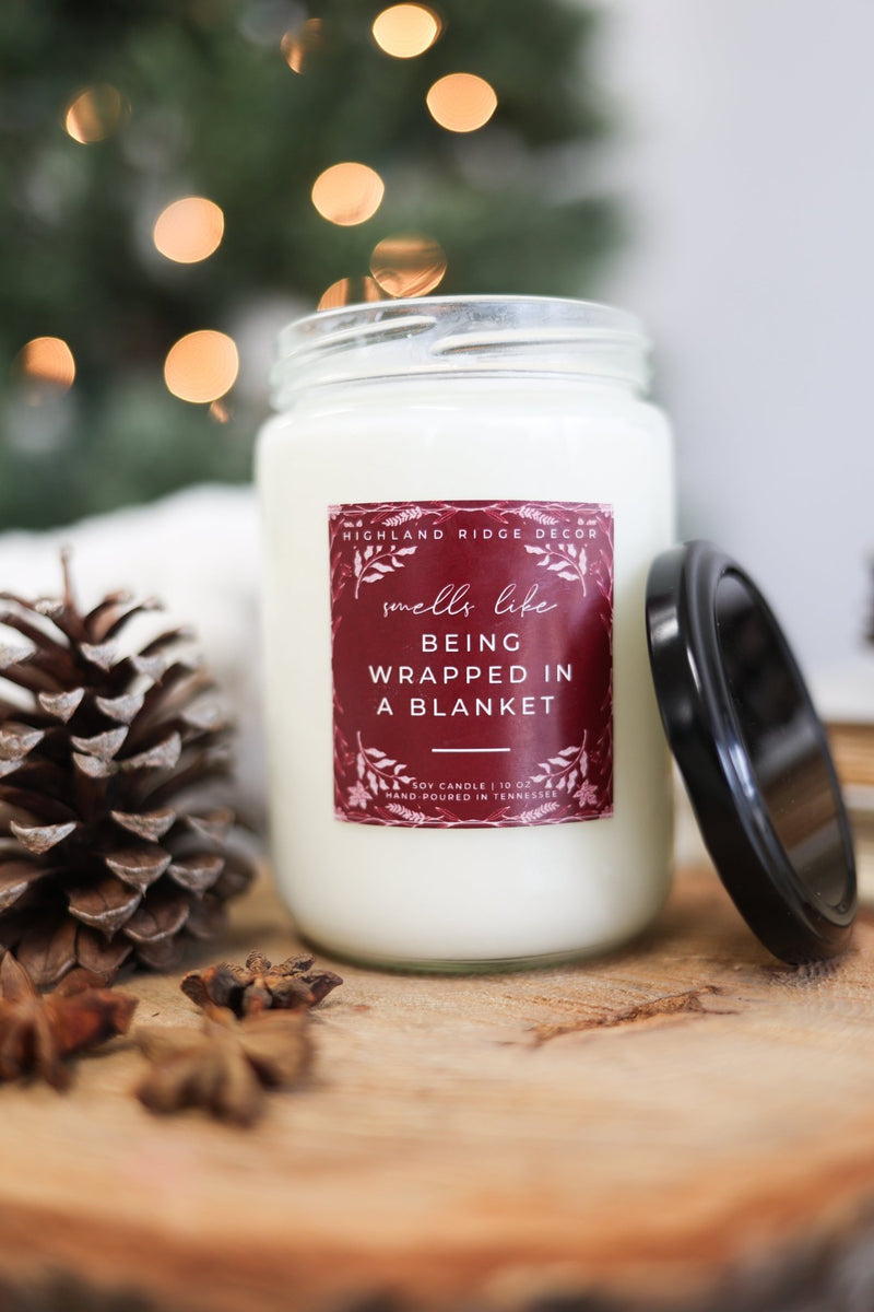 Being Wrapped In A Blanket Candle in a glass jar with holiday decor and pinecones.